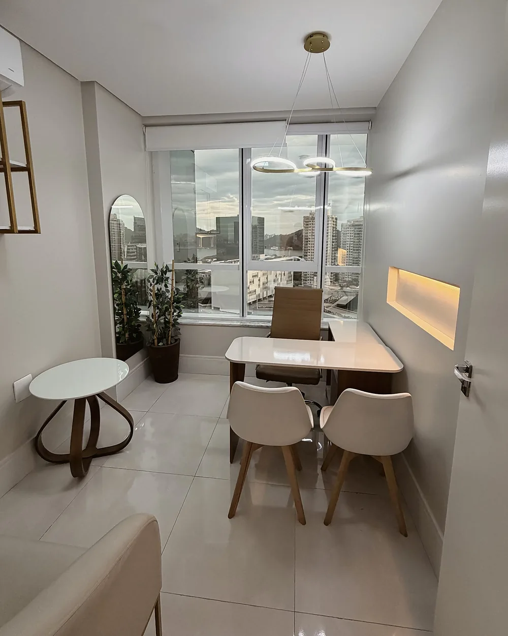 Sala de atendimento com mesa, duas cadeiras brancas, espelho em arco e janela para a cidade